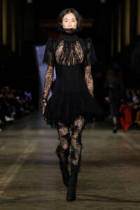 Model walks a fashion runway in a black lace Victorian-inspired dress with high collar, sheer bodice, and ruffled skirt, paired with lace tights and heeled boots