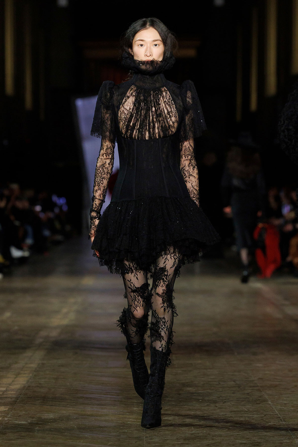 Model walks a fashion runway in a black lace Victorian-inspired dress with high collar, sheer bodice, and ruffled skirt, paired with lace tights and heeled boots