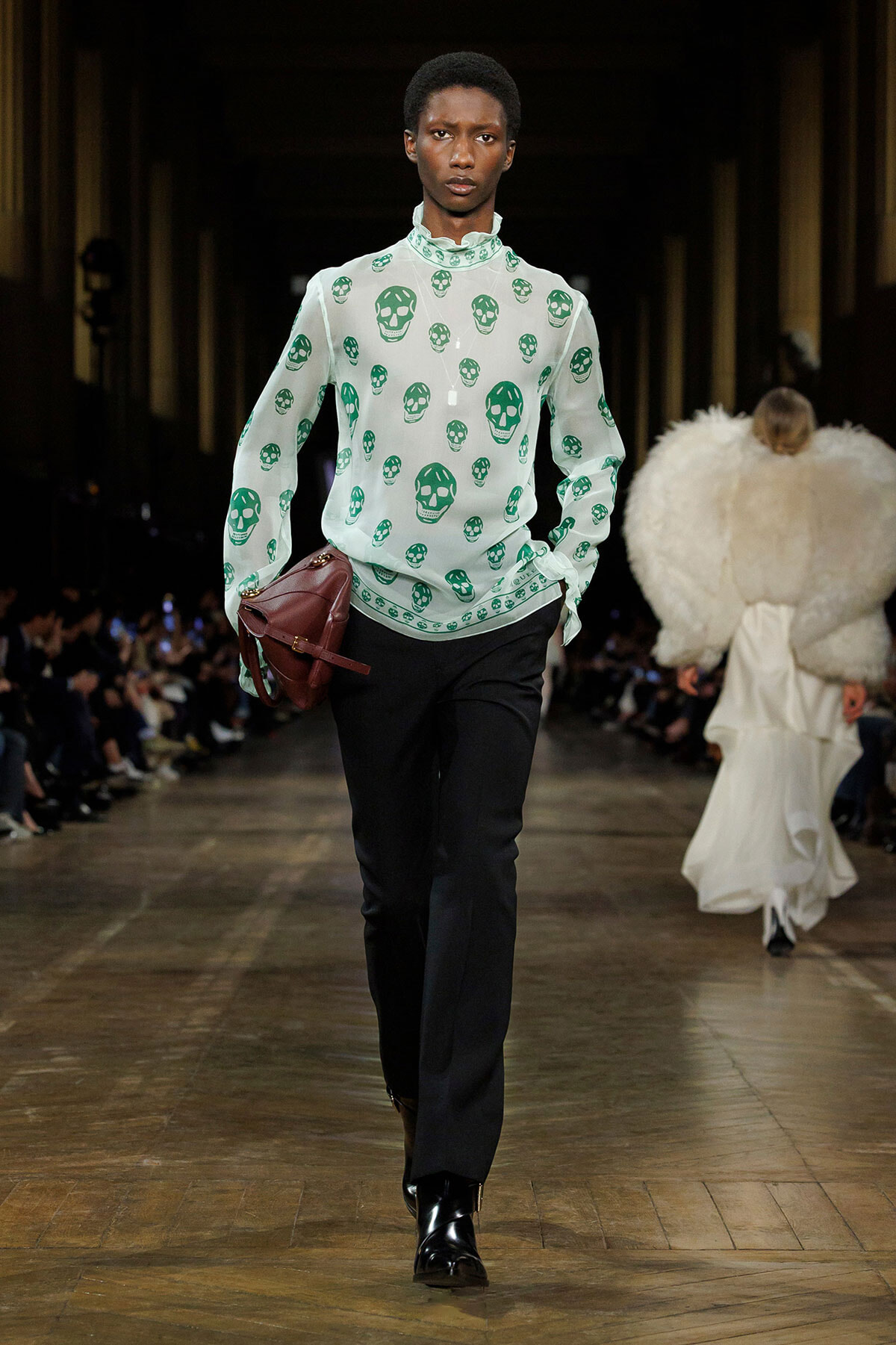 Male model walking a fashion runway in a mint-green skull-print blouse, black trousers, and a burgundy side bag.