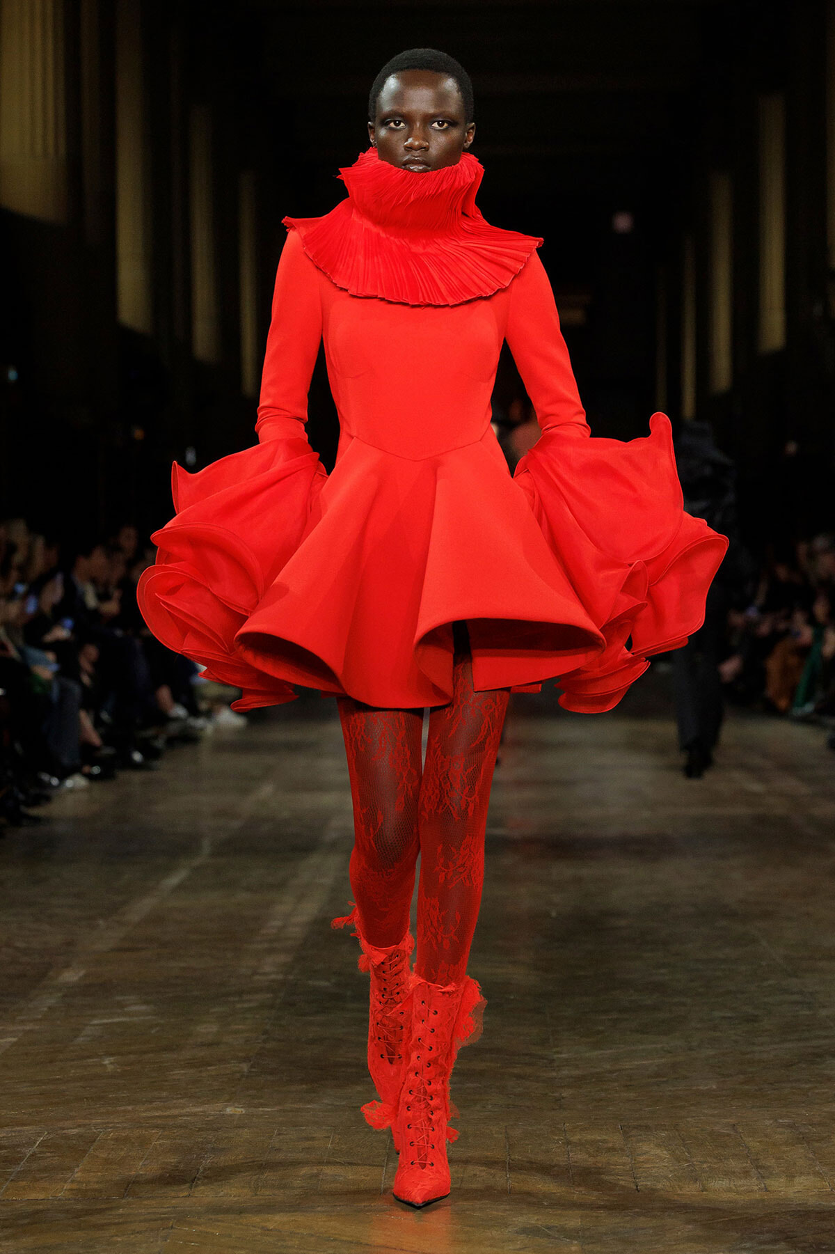 Model walks a fashion runway in a vivid red sculptural dress with an oversized ruffled collar and dramatic voluminous sleeves, paired with red lace tights and lace-up boots.