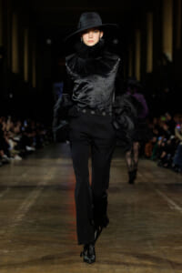 Model walking runway in an all-black velvet outfit with a high ruffled collar and wide-brim hat.