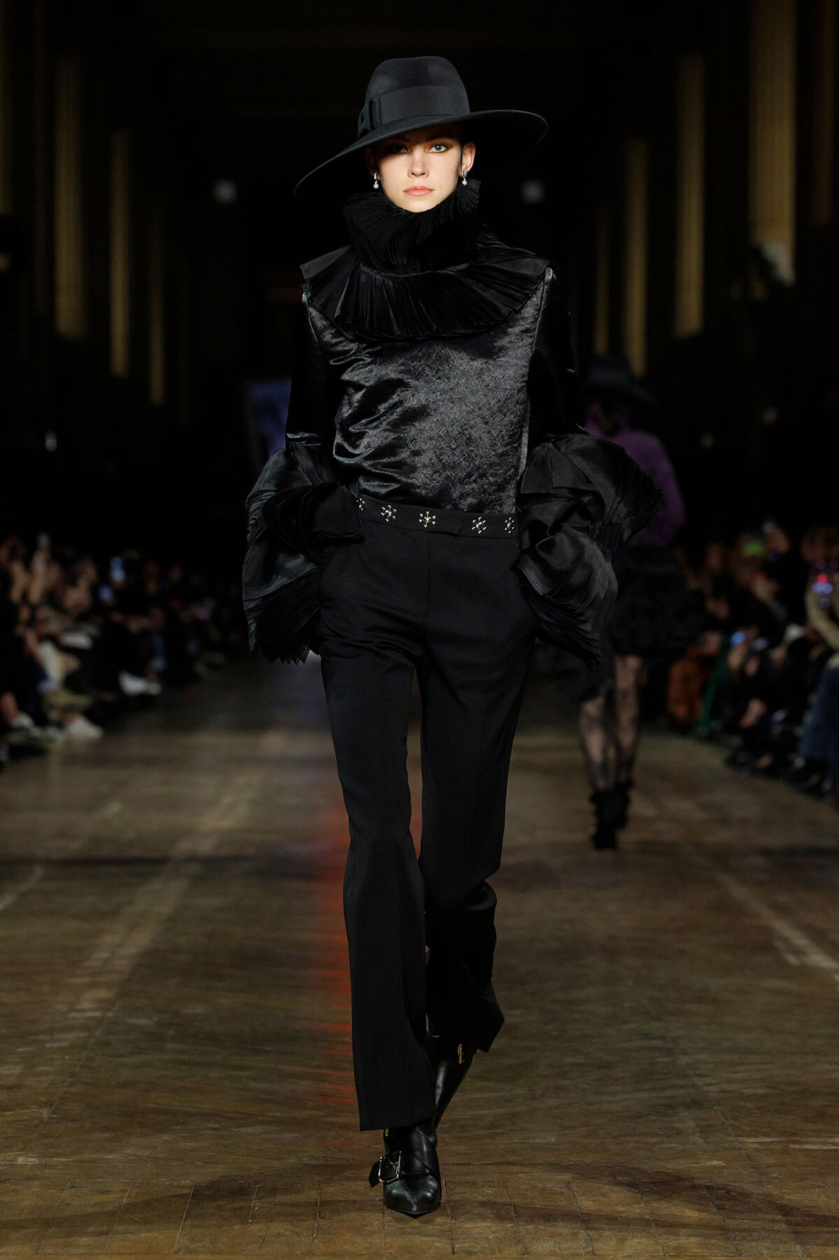Model walking runway in an all-black velvet outfit with a high ruffled collar and wide-brim hat.