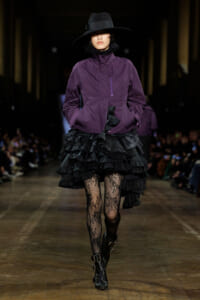 Model walking a runway in a purple zip-front jacket, a voluminous black layered skirt, lace tights, and black ankle boots with a wide-brimmed hat.