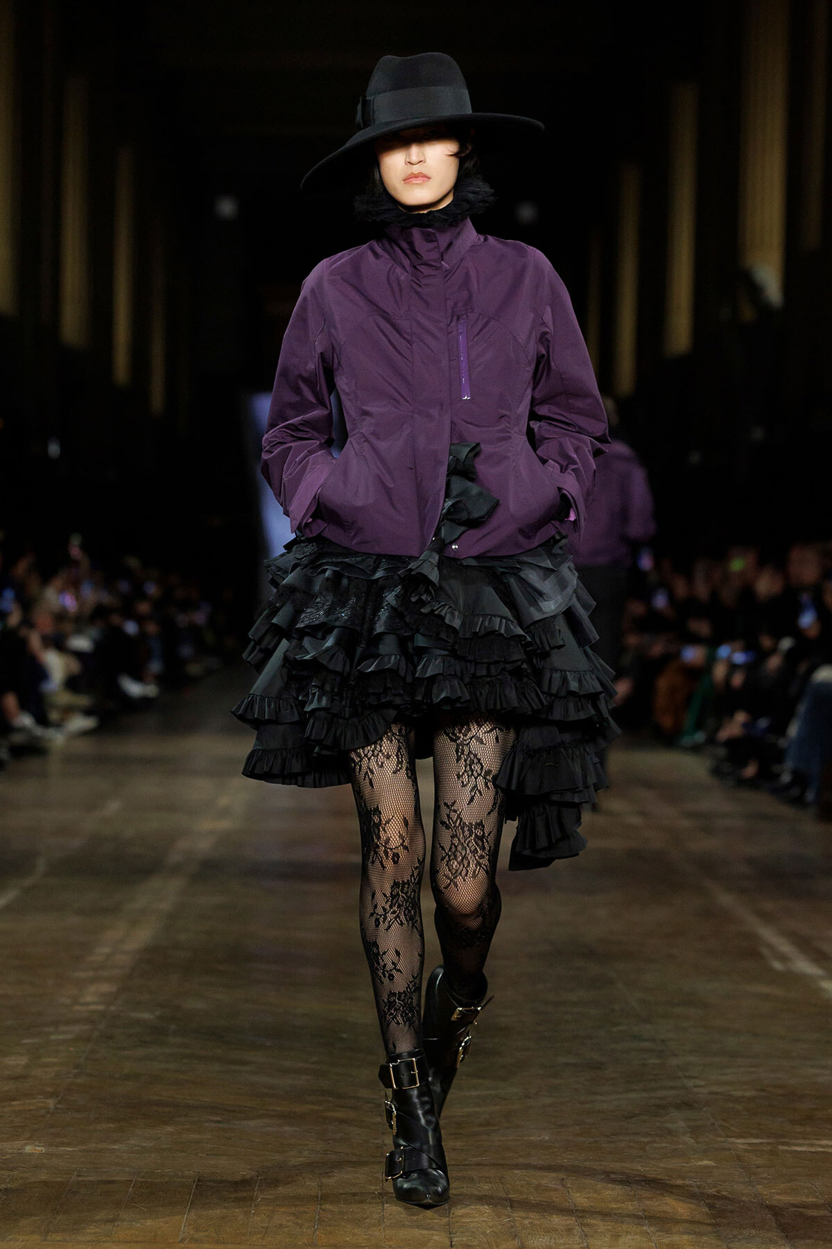 Model walking a runway in a purple zip-front jacket, a voluminous black layered skirt, lace tights, and black ankle boots with a wide-brimmed hat.