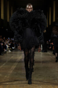 Model walks the runway in a dramatic oversized black faux-fur coat, paired with a sheer lace-topped dress and thigh-high boots.