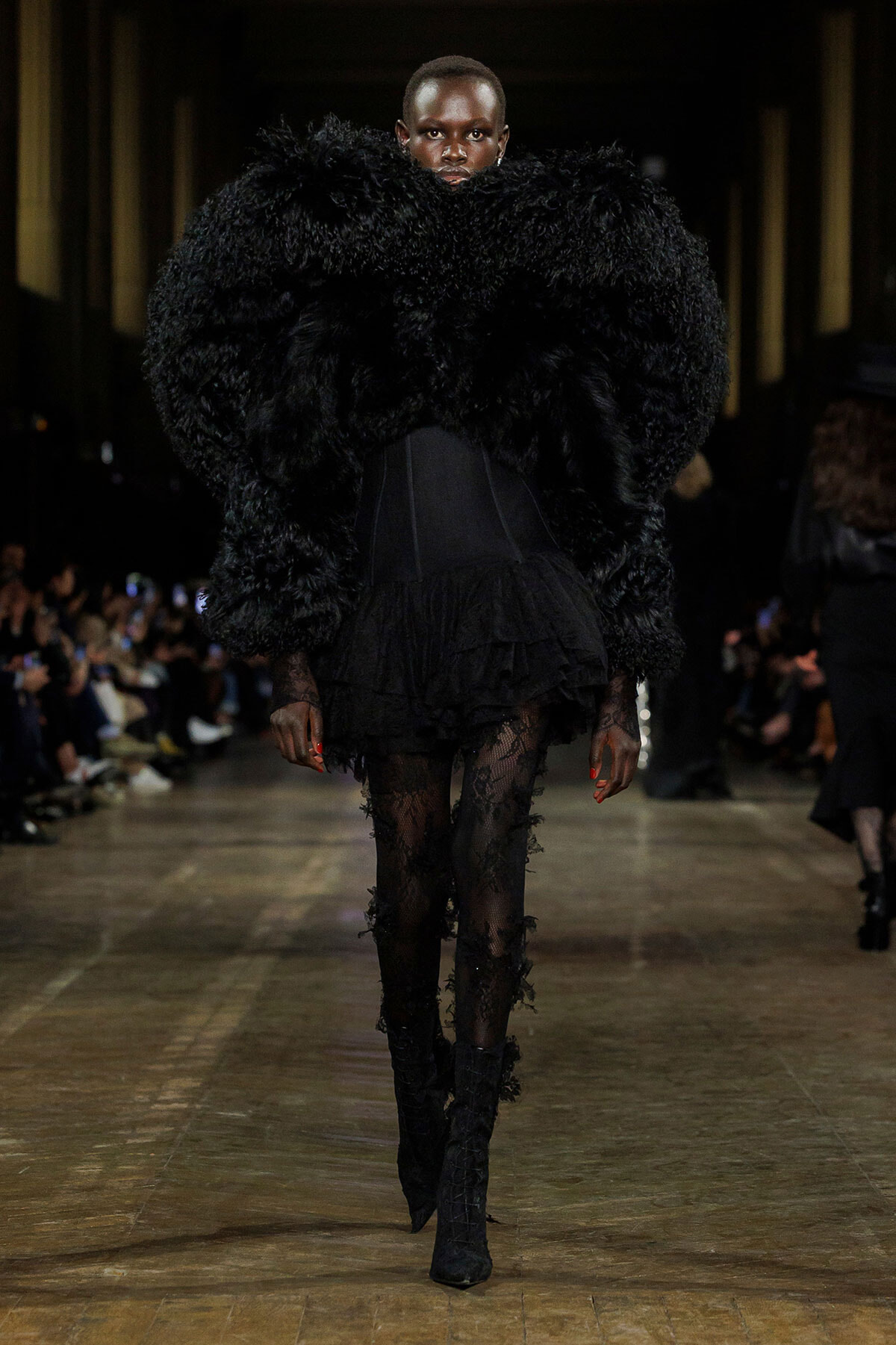 Model walks the runway in a dramatic oversized black faux-fur coat, paired with a sheer lace-topped dress and thigh-high boots.