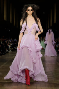 Model on a runway wearing a lavender pink gown with cascading ruffles, sheer bodice, sunglasses, and red boots.