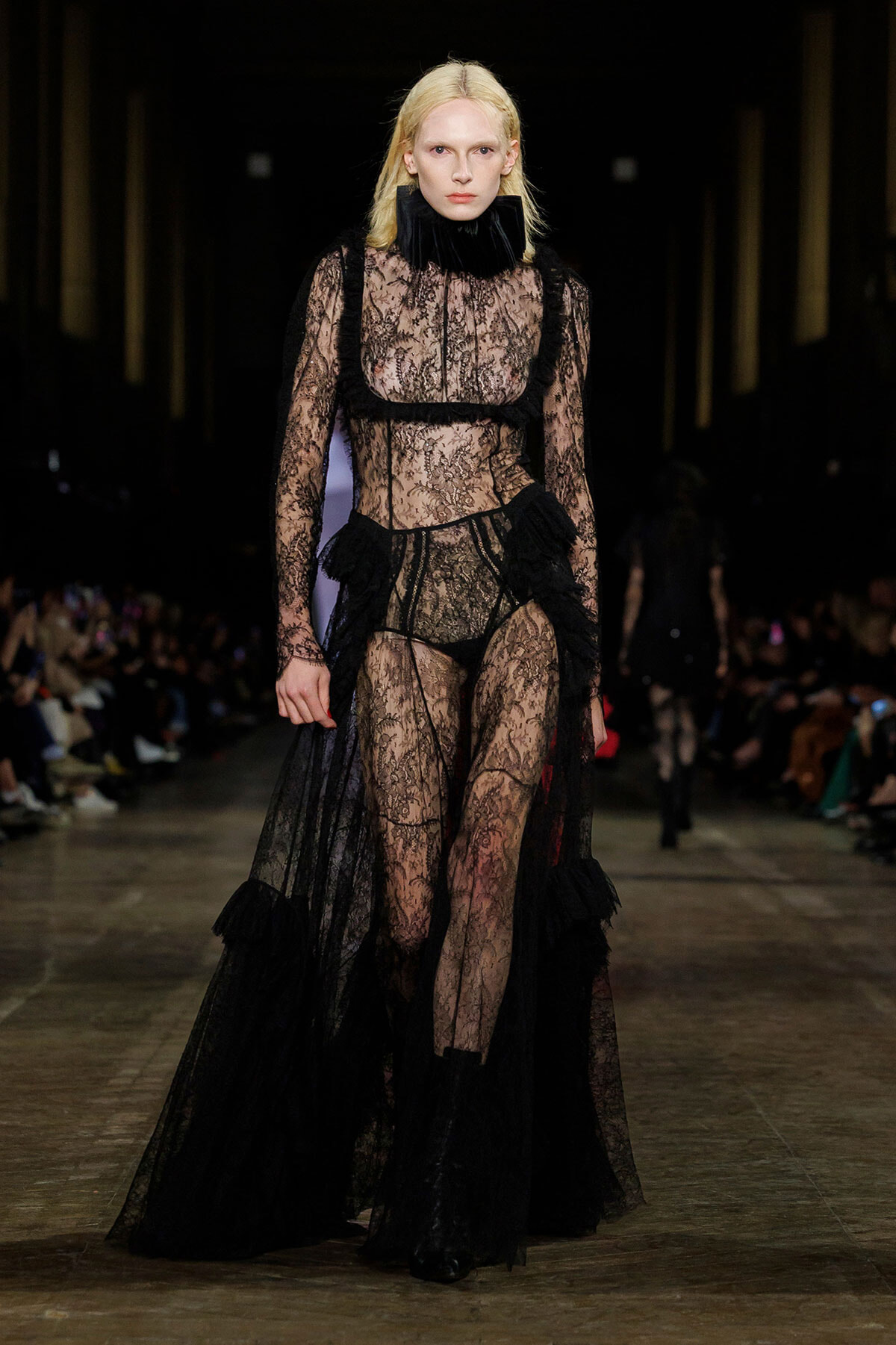 Model walking the runway in a black sheer lace gown with a high ruffled collar and long train, staged in a dark fashion show.