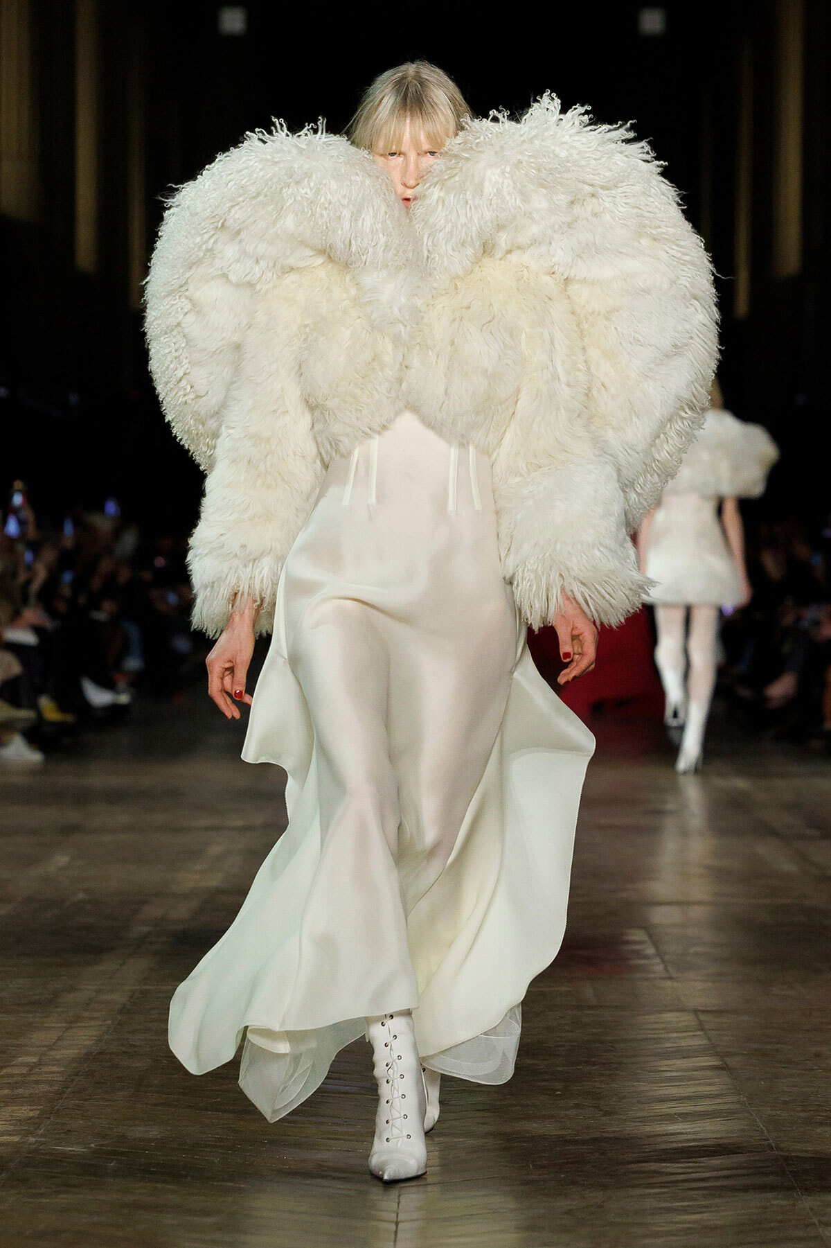 Model walking the runway in a white satin gown with an oversized fluffy white feather cape and white lace-up boots.