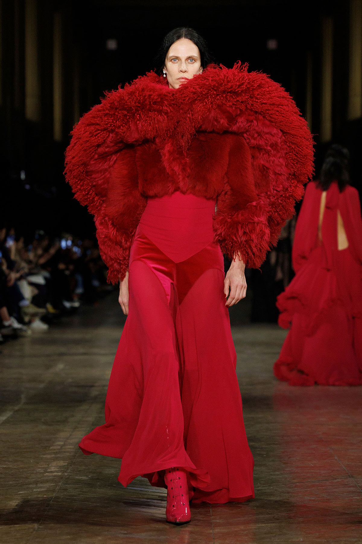 Model on a runway wearing a dramatic red feathered cape and a matching red gown with flowing skirt.