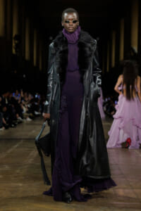 Model walks a fashion runway in a long black leather coat with a fur collar over a dark purple dress and matching turtleneck, wearing small sunglasses and carrying a black fringe bag.