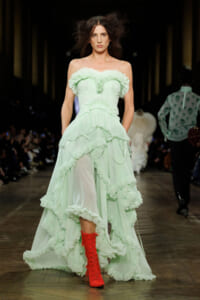 Model walking a runway in a pale mint green off-the-shoulder gown with layered ruffles and bright red lace-up boots on the catwalk.