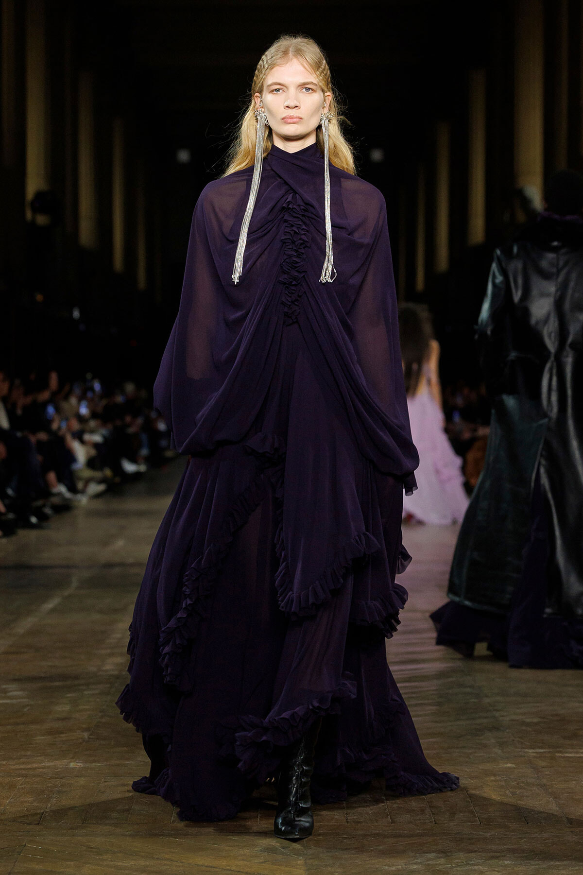 Model on a fashion runway wearing a deep purple, floor-length gown with dramatic draped ruffles and sheer, voluminous sleeves, accessorized with long silver earrings.