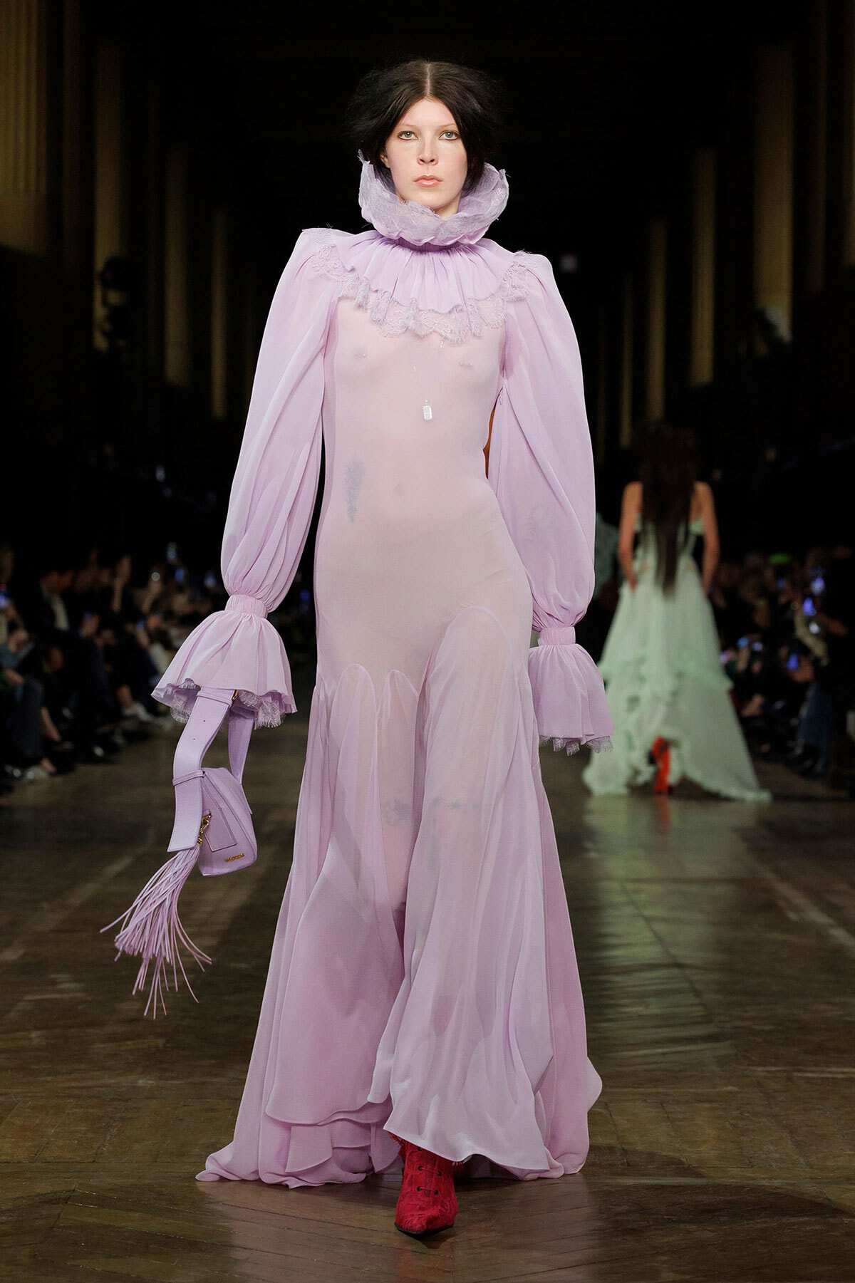 Model walks the runway in a sheer lavender gown with ruffled high collar and voluminous sleeves, holding a matching fringe purse.