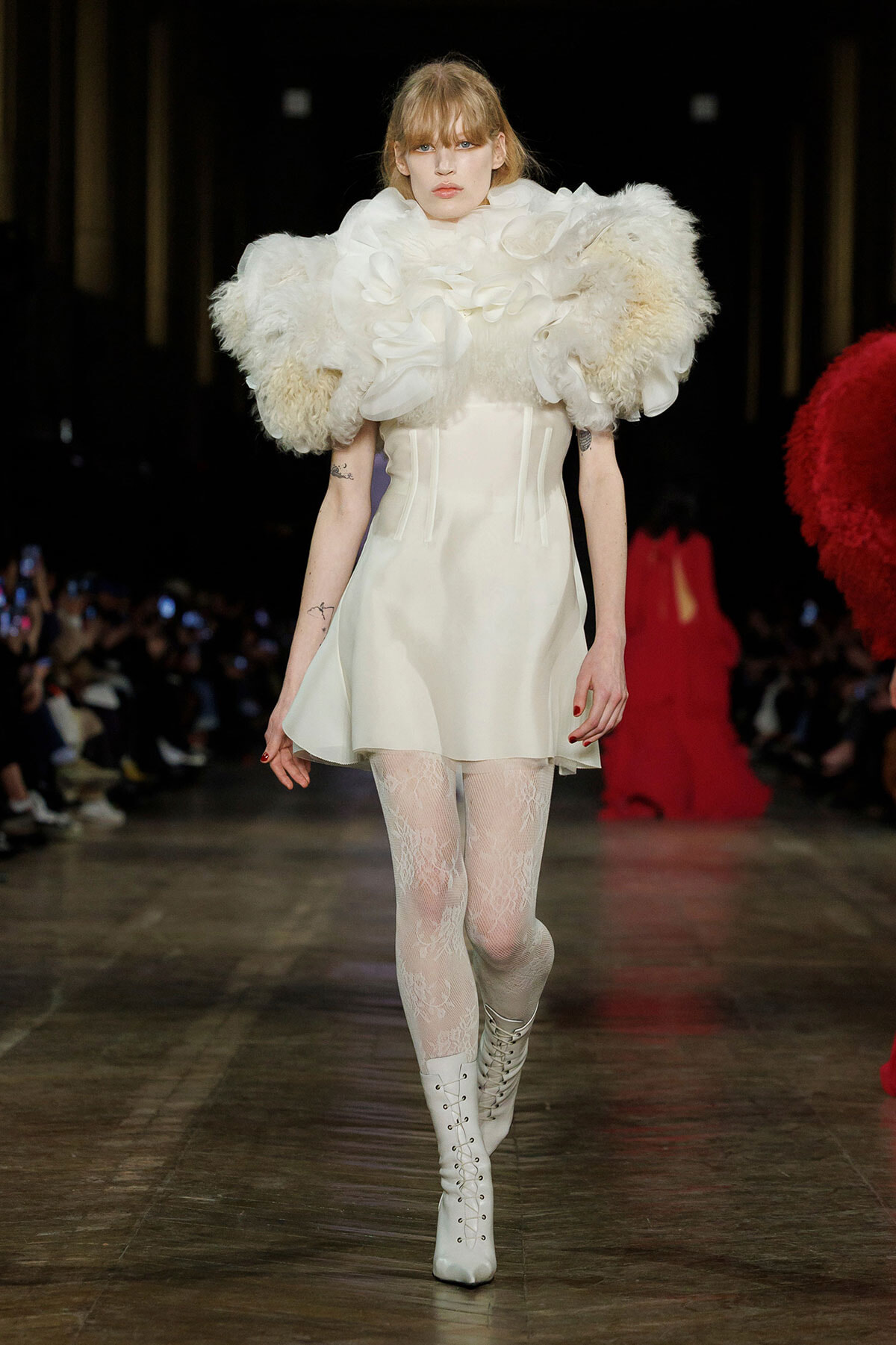Runway model in an ivory mini dress with dramatic fluffy feather shoulders, lace tights, and white lace-up boots.