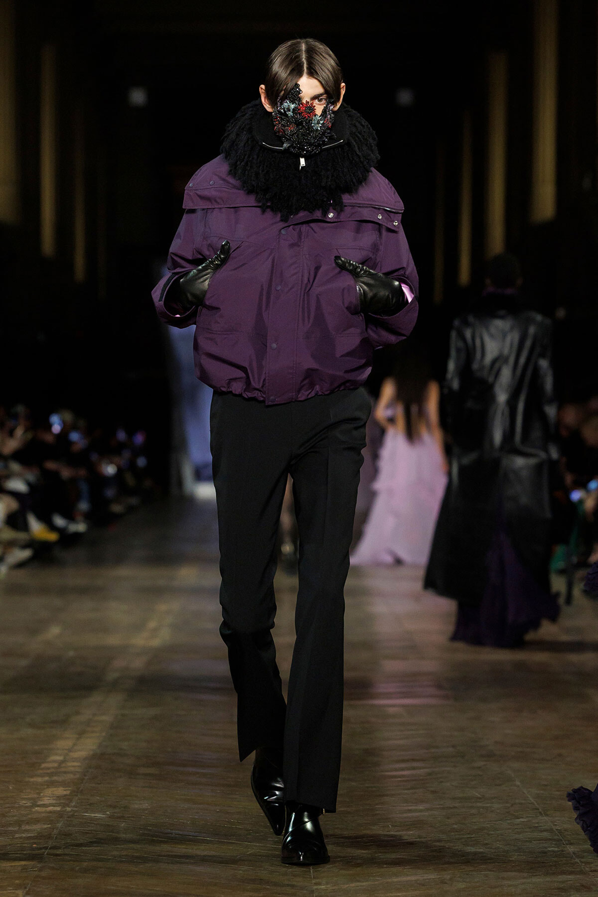 Runway model wearing a purple puffer jacket, black gloves, and black pants with a floral-faced mask, walking the catwalk.