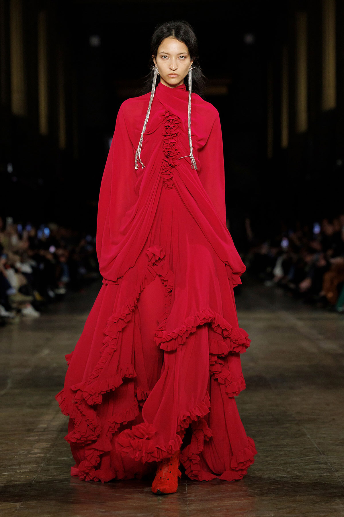 Model walking the runway in a bright red, floor-length gown with cascading ruffles and voluminous sleeves, wearing long silver earrings.