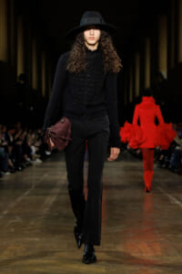 Model walking a fashion runway in an all-black ensemble: wide-brim hat, textured cardigan, black trousers, and a burgundy shoulder bag.
