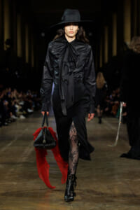 Model walks a runway in an all-black ensemble: wide-brim hat, ruffled blouse, tailored skirt with a lace tights slit, ankle boots, and a black handbag with a red scarf.