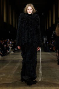 Model walks the runway in a long, black, textured fur coat, holding a silver fringe accessory at her side.