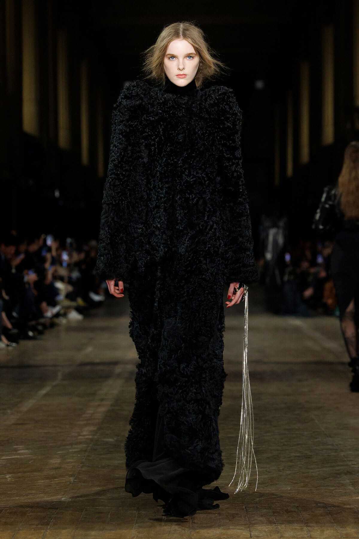 Model walks the runway in a long, black, textured fur coat, holding a silver fringe accessory at her side.
