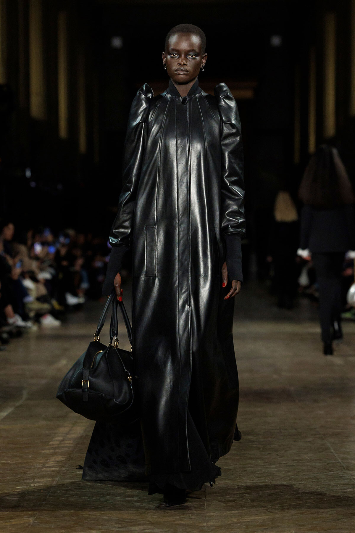 Model on a fashion runway wearing a floor-length glossy black leather dress with exaggerated shoulders, carrying a black handbag.