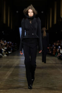 Model walking the runway in a black double-breasted tailored blazer with matching black trousers and a lace top underneath, completing a sleek monochrome look.