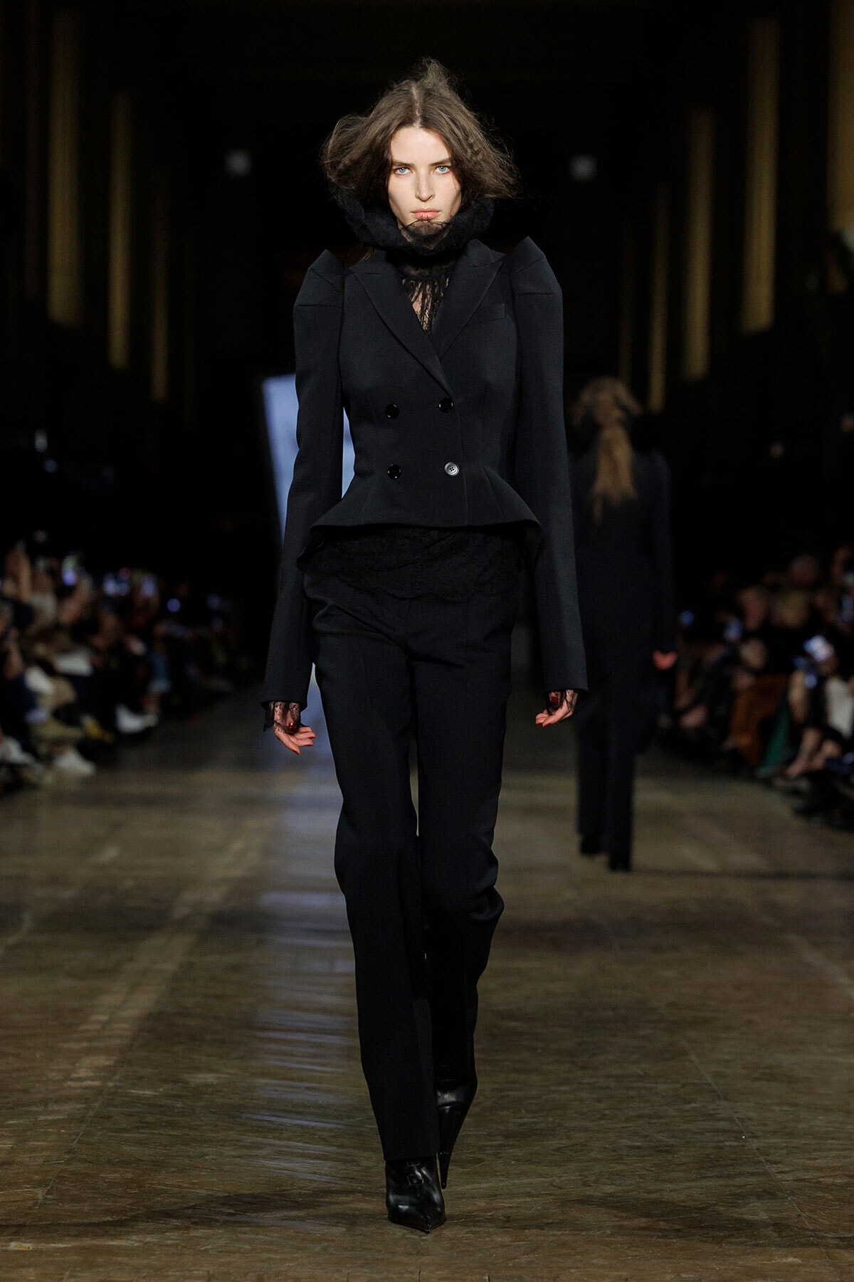 Model walking the runway in a black double-breasted tailored blazer with matching black trousers and a lace top underneath, completing a sleek monochrome look.