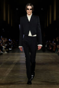 Male model walks a fashion runway in a black pinstripe three-piece suit with a cropped blazer, white ruffled shirt, and black boots, wearing sunglasses.