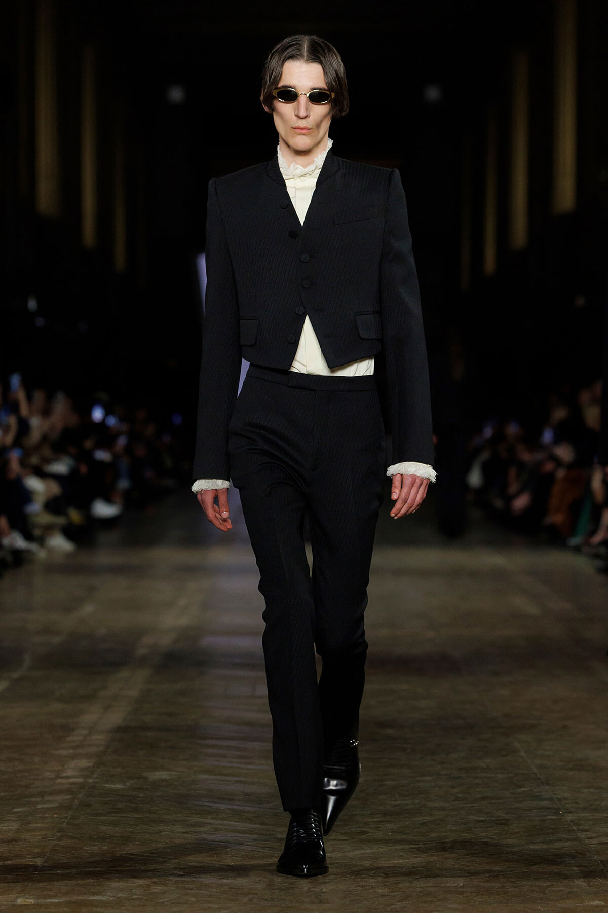 Male model walks a fashion runway in a black pinstripe three-piece suit with a cropped blazer, white ruffled shirt, and black boots, wearing sunglasses.