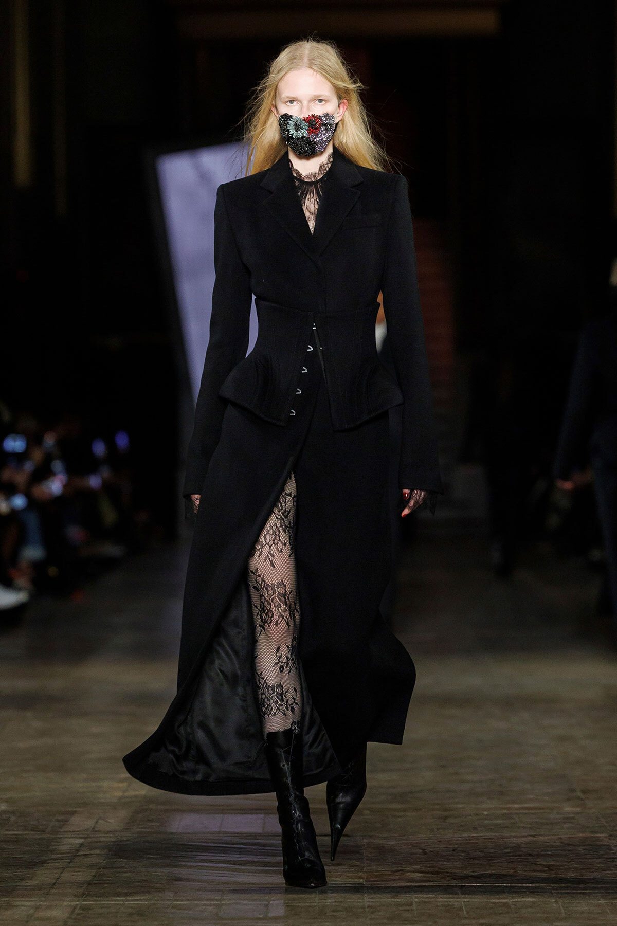 Model walks a dark, tailored black coat with cinched waist and lace tights on a runway, wearing a floral mask.