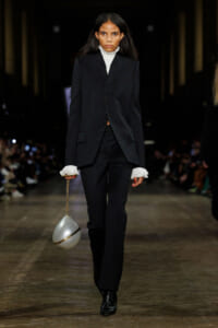 Female model walking a fashion runway in a black tailored suit with a white high-neck top and ruffled cuffs, carrying a silver spherical handbag on a gold chain.