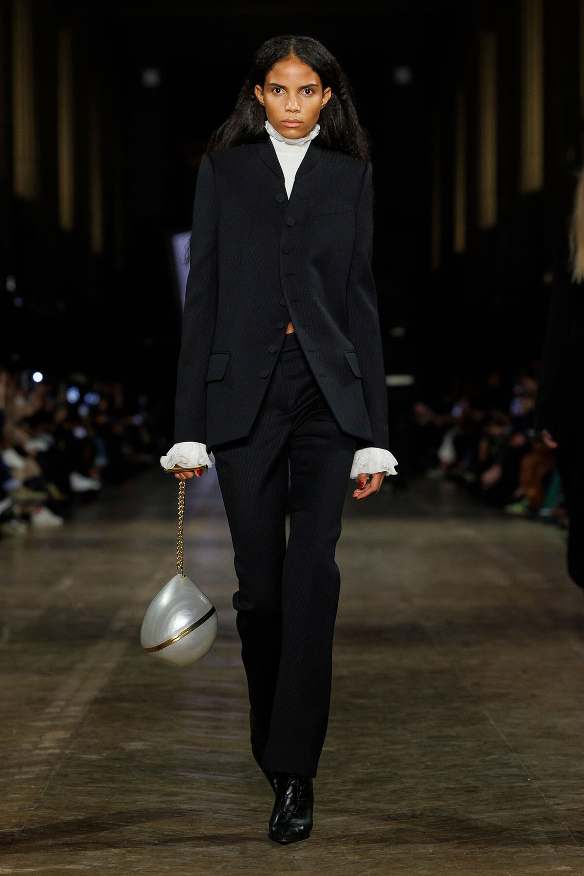 Female model walking a fashion runway in a black tailored suit with a white high-neck top and ruffled cuffs, carrying a silver spherical handbag on a gold chain.