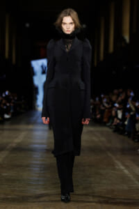 Female model walks a fashion runway in a long black tailored coat with structured shoulders and a lace high-neck underneath, black boots, and minimal makeup amid a dimly lit backstage-like setting.