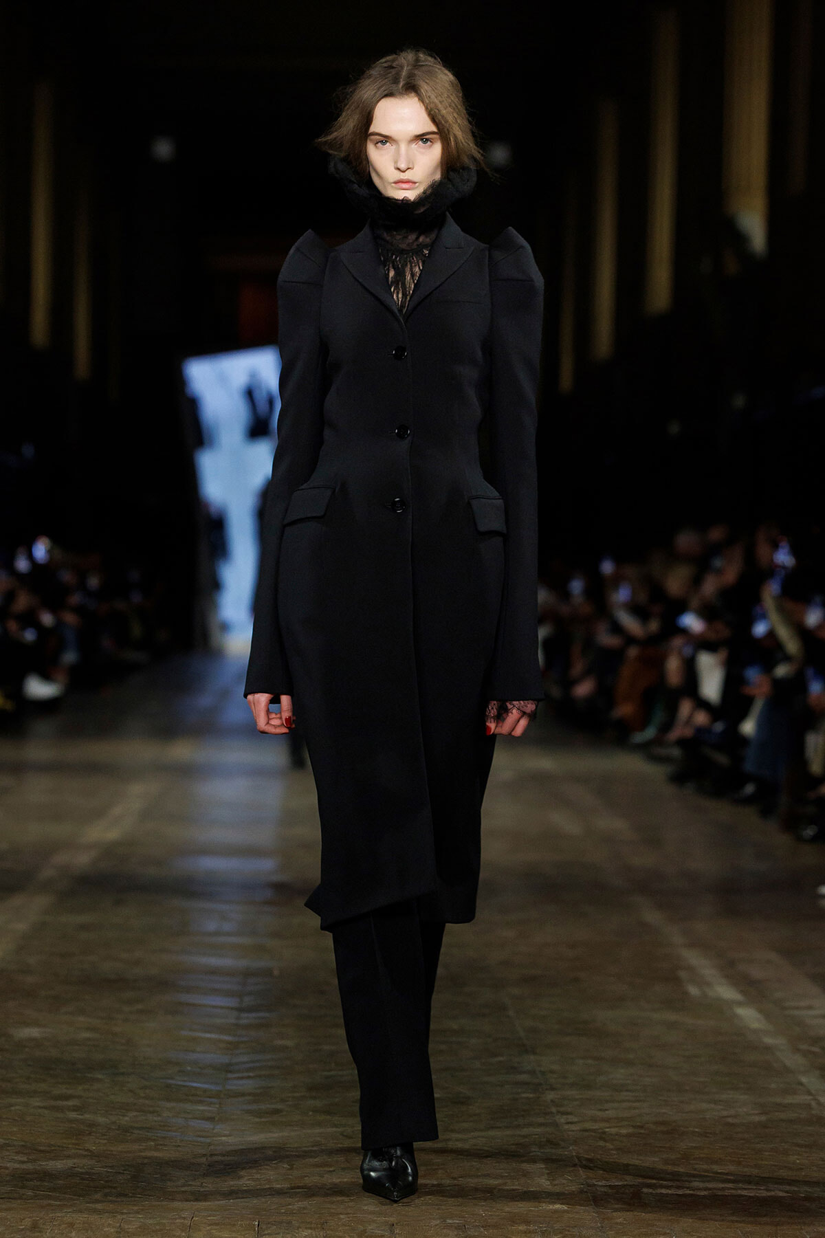 Female model walks a fashion runway in a long black tailored coat with structured shoulders and a lace high-neck underneath, black boots, and minimal makeup amid a dimly lit backstage-like setting.