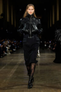Model on a runway wearing a glossy black leather jacket with buckle straps, a black midi skirt, and patterned lace tights paired with buckled ankle boots.
