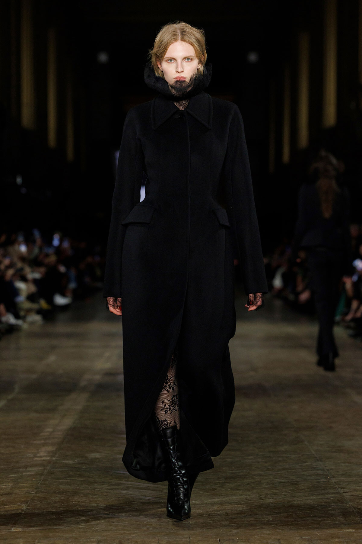 Model walks a dark, tailored black coat with lace high collar, black gloves, and lace tights under knee-high boots on a runway.