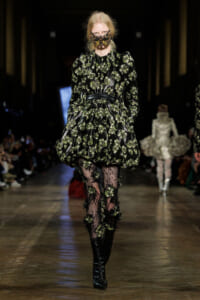 Model walks the runway in a black-green floral puff-sleeve dress with a cinched waist, lace tights, and knee-high boots paired with a jeweled mask.
