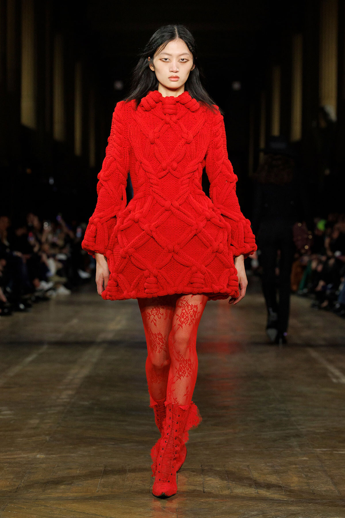 Model walking the runway in a bold red oversized knitted coat with a chunky cable pattern, paired with red lace tights and boots?