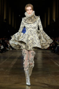 Model on a runway wearing an ivory floral brocade dress with a large pleated ruff collar and layered voluminous skirt, paired with lace tights and metallic boots.