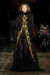 Model walks the runway in a black coat with gold embroidered vines running along the front and cuffs, revealing sheer black pants underneath.