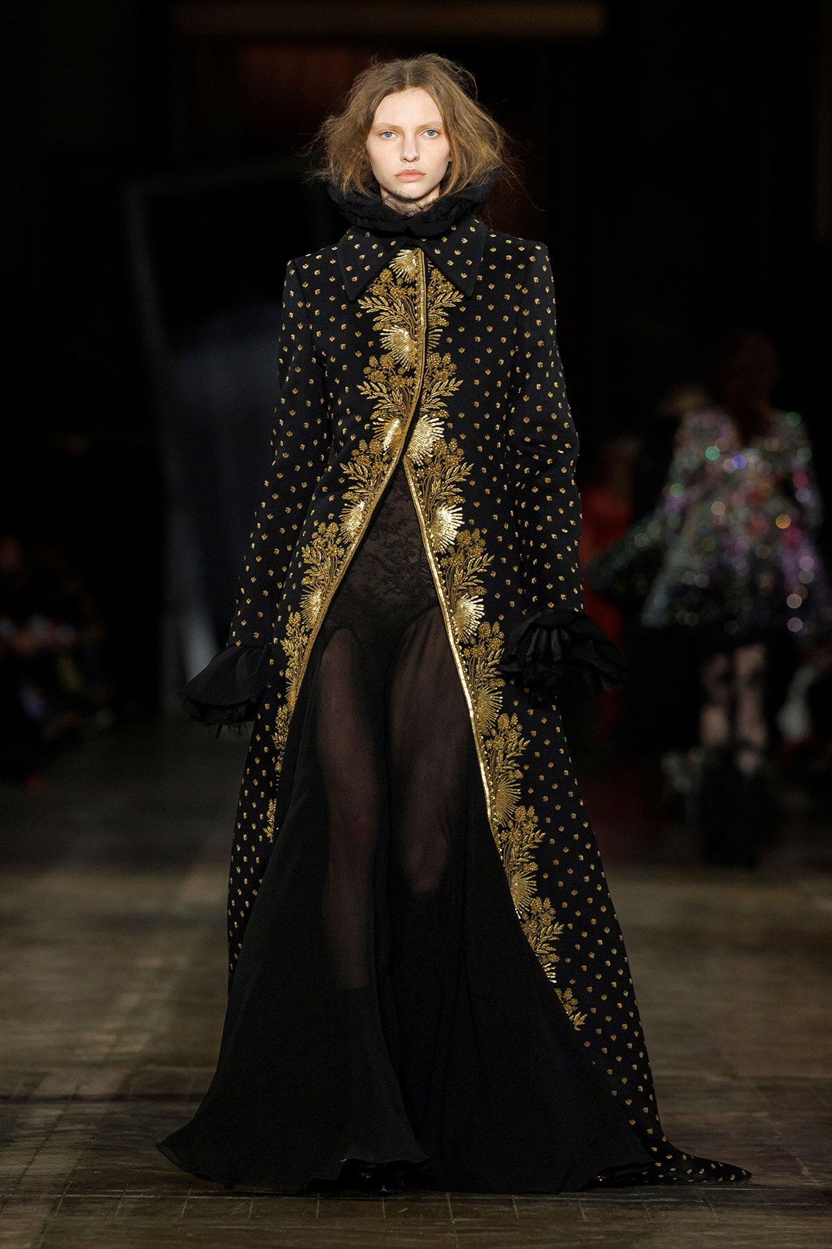 Model walks the runway in a black coat with gold embroidered vines running along the front and cuffs, revealing sheer black pants underneath.