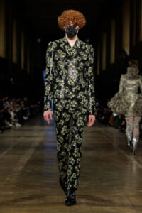 Model on a runway wearing a black floral-print suit with green leaves and a matching face mask.