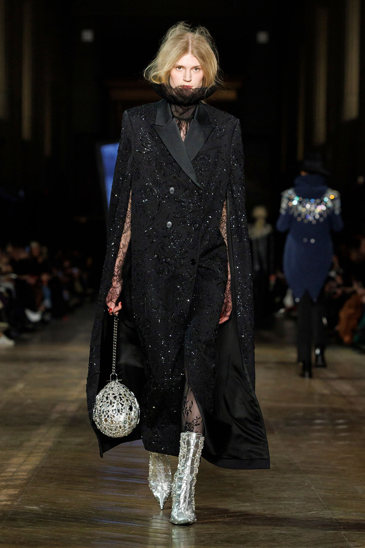 Model on a runway wearing a black sequined double-breasted cape coat with sheer lace sleeves and a sheer high collar, paired with sparkly silver boots and a jeweled spherical handbag.