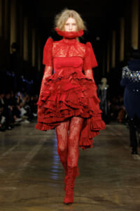Model on a runway wearing a red lace dress with layered ruffles, high collar and matching tights and boots.