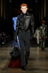 Male fashion model with short red hair walks the runway in a black embossed leather jacket, dark trousers, black gloves, and carries a black handbag.