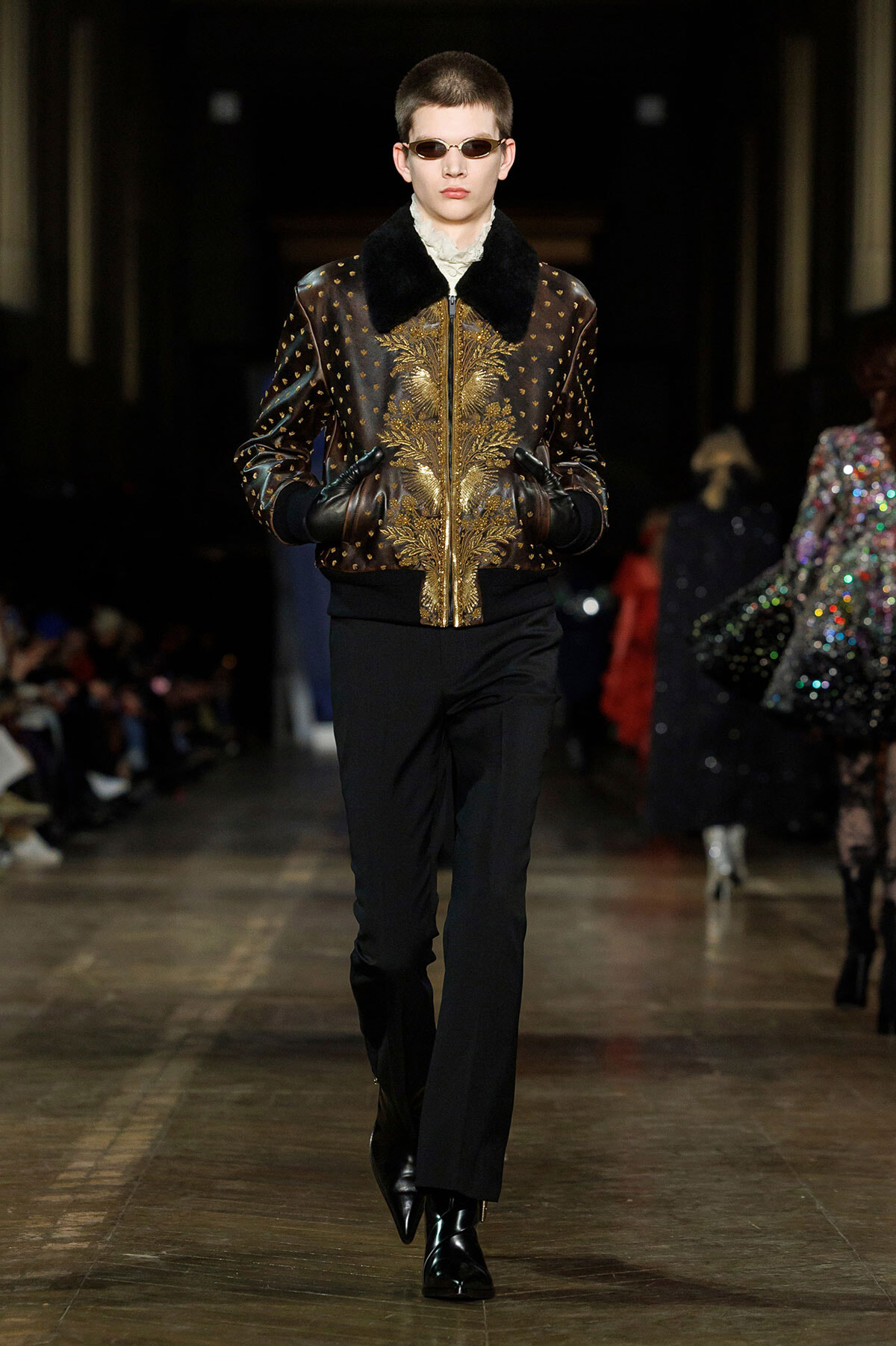 Model walking the runway in a black leather bomber jacket with gold embroidery and a black fur collar, paired with black pants, gloves, and shiny black boots, wearing sunglasses.