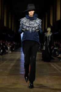 Model on a runway wearing a navy blue embroidered top with silver beadwork, black gloves, and a wide-brim hat.