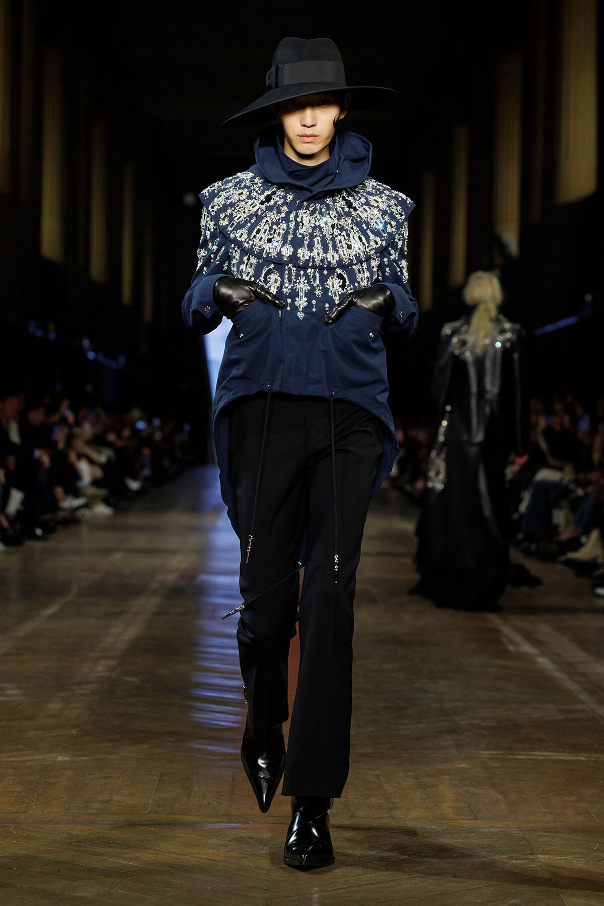 Model on a runway wearing a navy blue embroidered top with silver beadwork, black gloves, and a wide-brim hat.