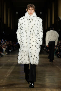 Model walks the runway in a long white fur coat with black feather-like markings, paired with black trousers and glossy boots.